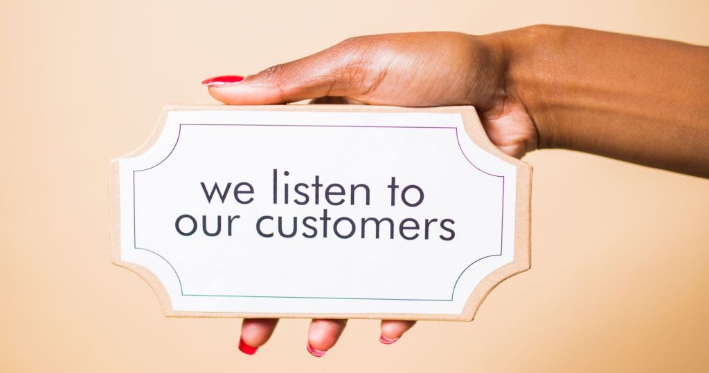A hand holding a sign saying 'we listen to our customers' on a neutral background.